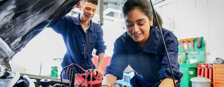 A woman mechanic looks into the hood of a car while a male colleague watches and both are smiling