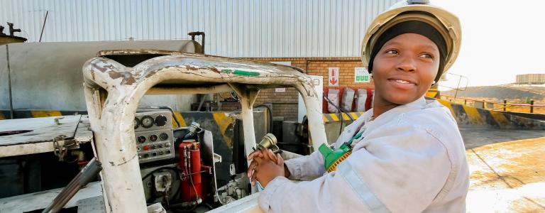 A female mine worker in South Africa operates machinery outside while smiling and looking off to the right