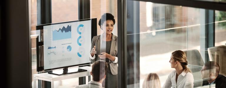 A woman in a business outfit presents to a group, as seen through a window