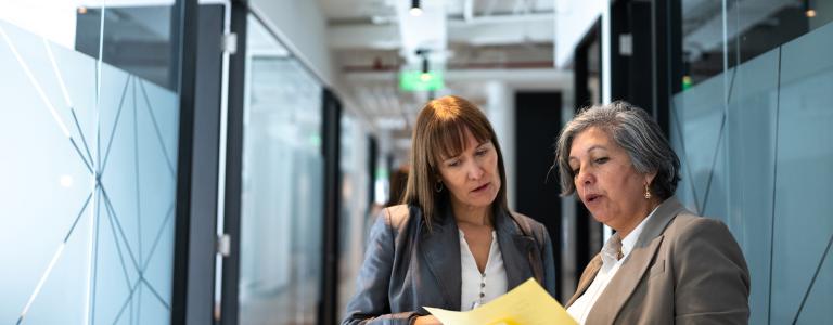 Two women in a hallway discussing business-related matters while examining a written document.