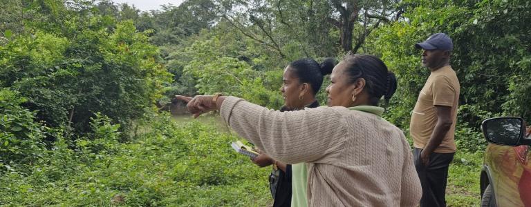 two women standing in a forested area, one pointing a finger