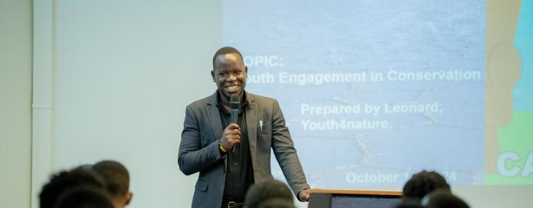 A young man holds a mic and stands in front of a small audience