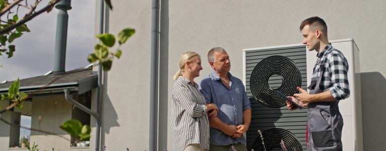 A technician uses a tablet to demonstrate heat pump features to a couple outdoors.