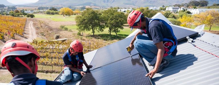 Three workers install solar panels on top a steel roof. There are trees and mountains in the background.