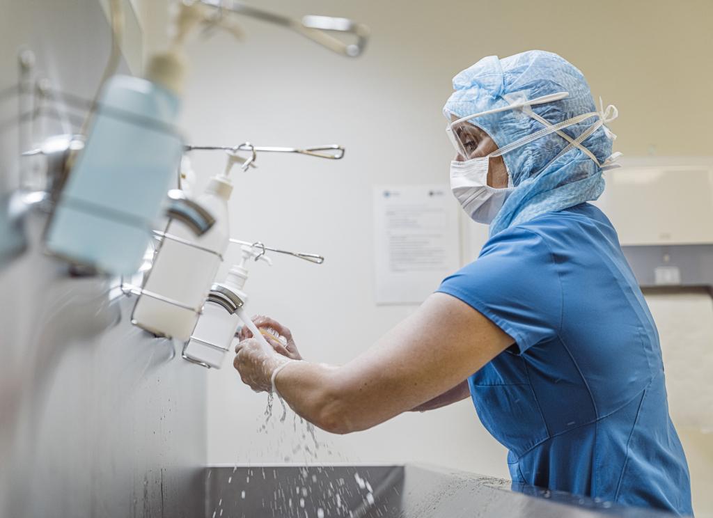 A woman in scrubs, mask and hairnet washes hands with soap over a sink