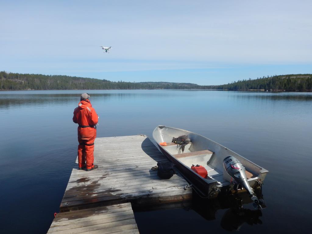 A man in an orange jumpsuit stands on a dock flying a drone with a boat on a lake next to him